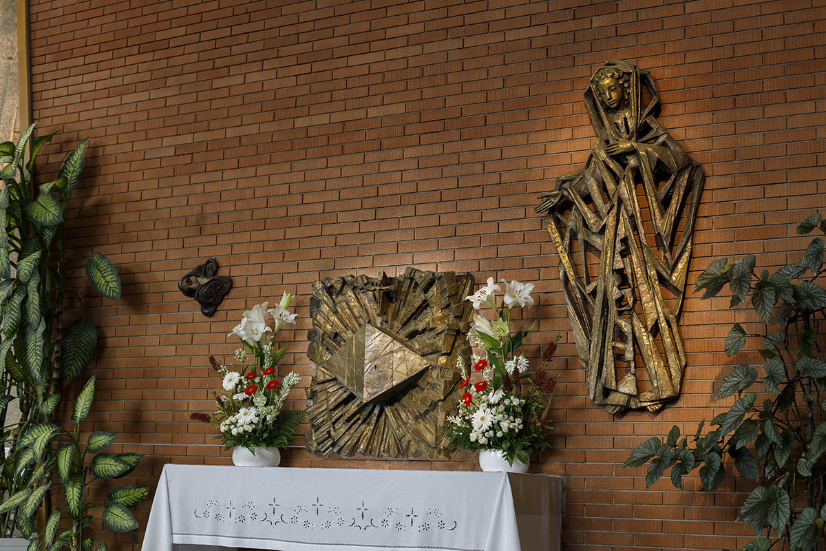 No altar, as figuras douradas sobre a parede de tijolo vigiam em silêncio. Arranjos florais e plantas completam o cenário onde tudo está prestes a acontecer.