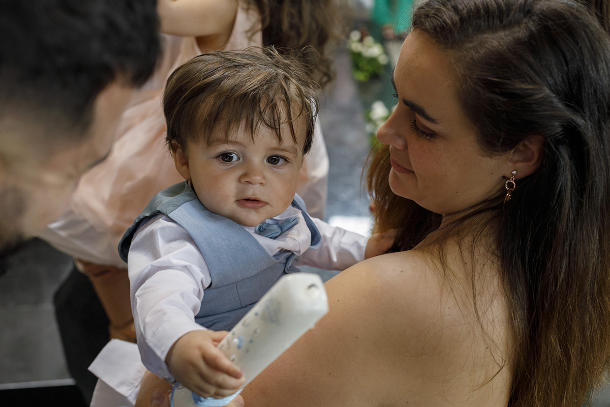 A mãe segura Gabriel nos braços. Ele contempla a vela do Batismo com seriedade, enquanto ela o observa com olhar terno. Um momento de entrega e amor.