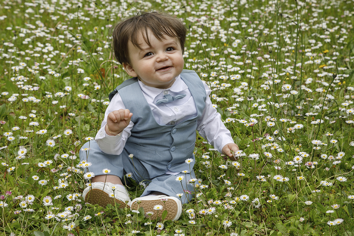Sobre um tapete de erva com florezinhas brancas, Gabriel está sentado ao centro. Com uma mão no chão e a outra no joelho, olha para a frente com alegria pura.