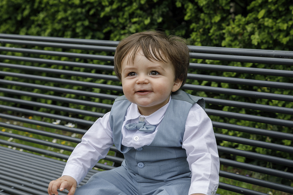 O pequeno Lourenço, sentado num banco cinzento, sorri com os olhos e o coração. A câmara capta-o da cintura para cima, num instante cheio de emoção e luz.
