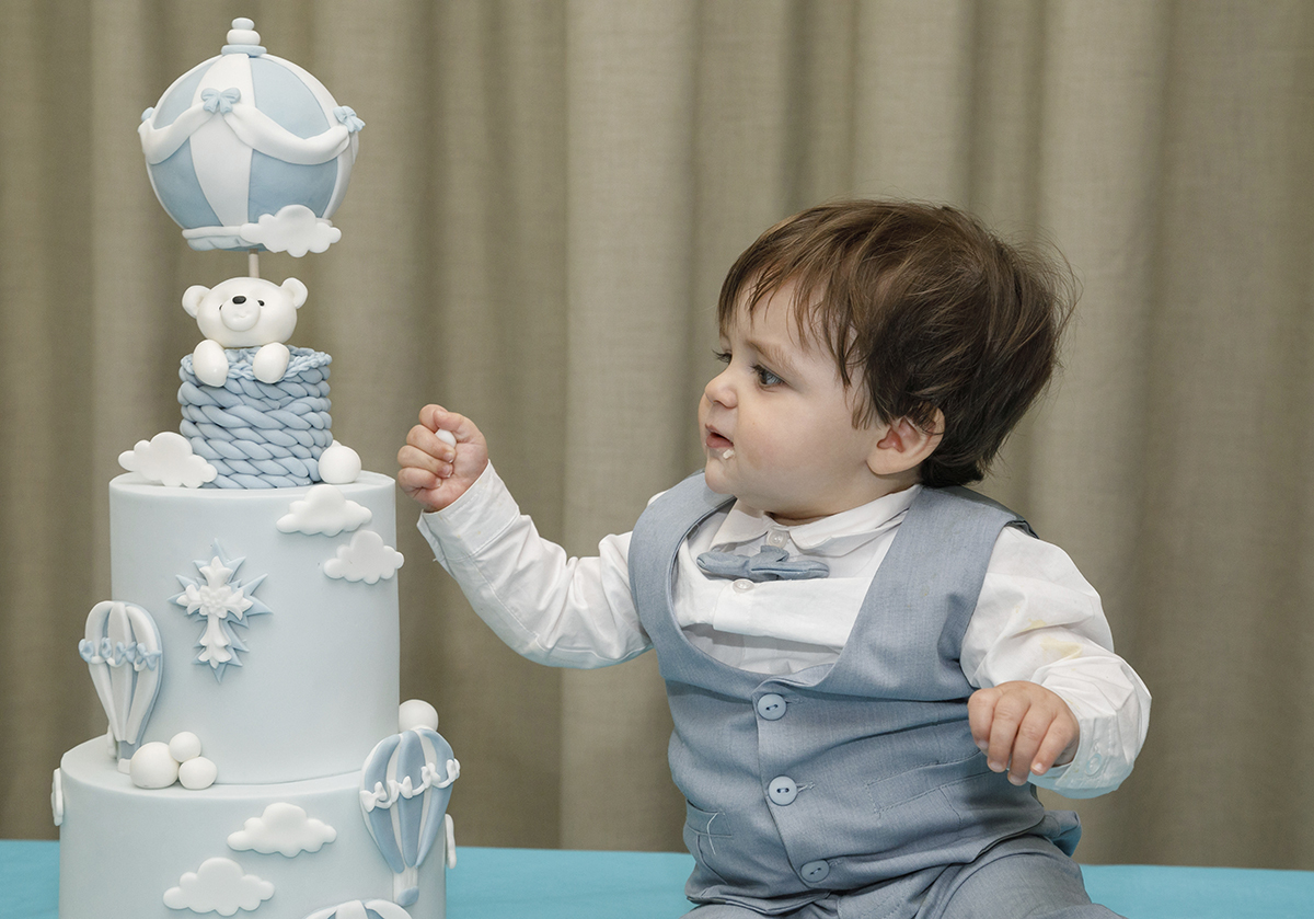 Junto ao seu bolo de dois andares em azul e branco, Gabriel quase toca na cobertura com curiosidade. O fundo é simples, com uma cortina bege, e a cena respira doçura e festa.