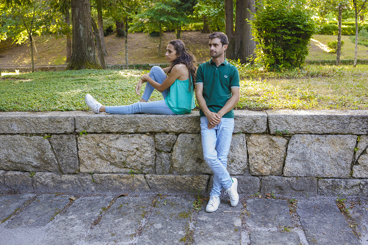 Diana senta-se no muro com pose descontraída, enquanto André permanece junto dela, sereno. Ambos olham para o horizonte, unidos por uma calma silenciosa.
