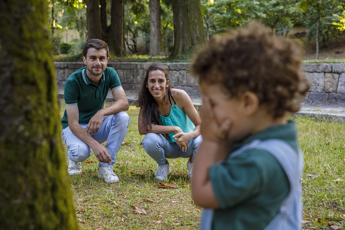 Tomé tapa o rosto com as mãos enquanto os pais, ao fundo, o observam sorridentes, talvez a contar para o jogo das escondidas.