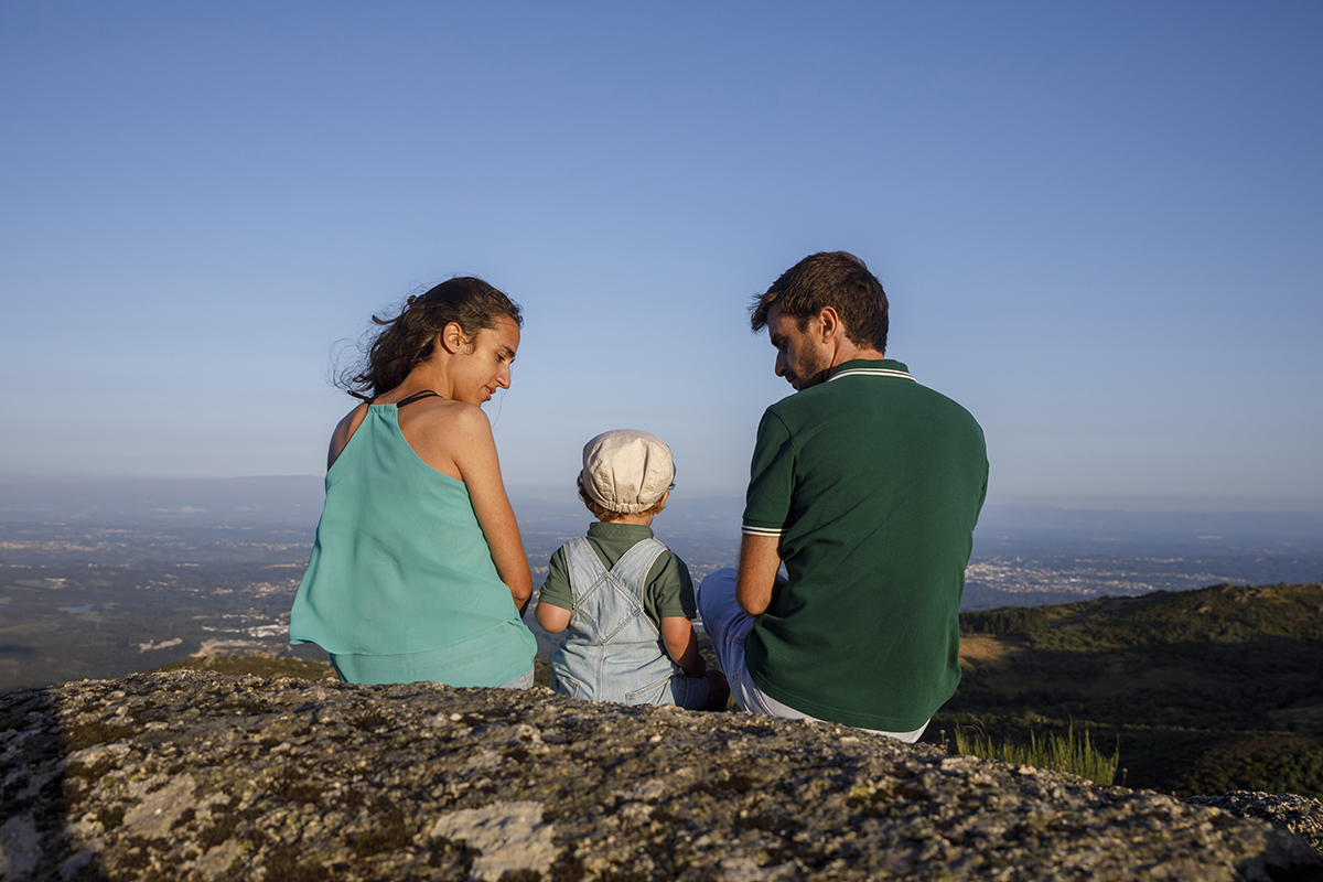 Sentados juntos no alto de uma rocha, Diana, André e Tomé contemplam a vista. Os pais olham o filho com serenidade, num cenário de céu, montanhas e amor.