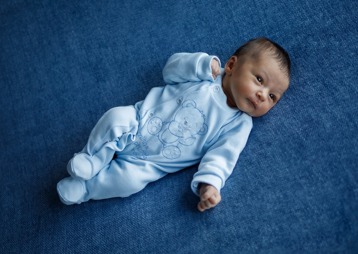 Deitado sobre um fundo azul suave, o pequeno Daniel descansa com um pijaminha azul encantador. Vemos o seu perfil delicado, com a cabeça virada para a direita, uma mãozinha perto do rosto e um olhar sonolento que derrete corações.