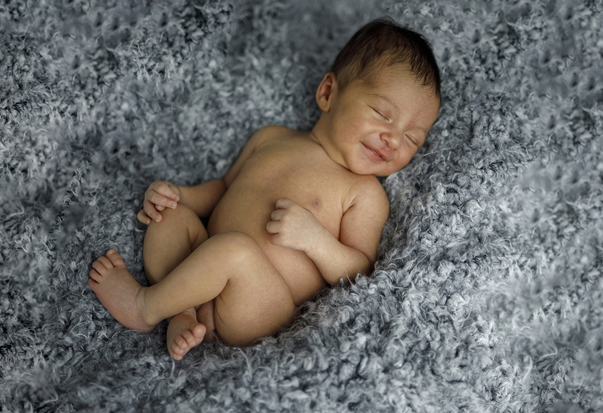 Deitado sobre uma manta branca e cinza fofa, Daniel sorri com os olhos fechados, numa expressão de sonho encantado. As pernas cruzadas e as mãozinhas repousadas compõem um retrato de paz e felicidade.