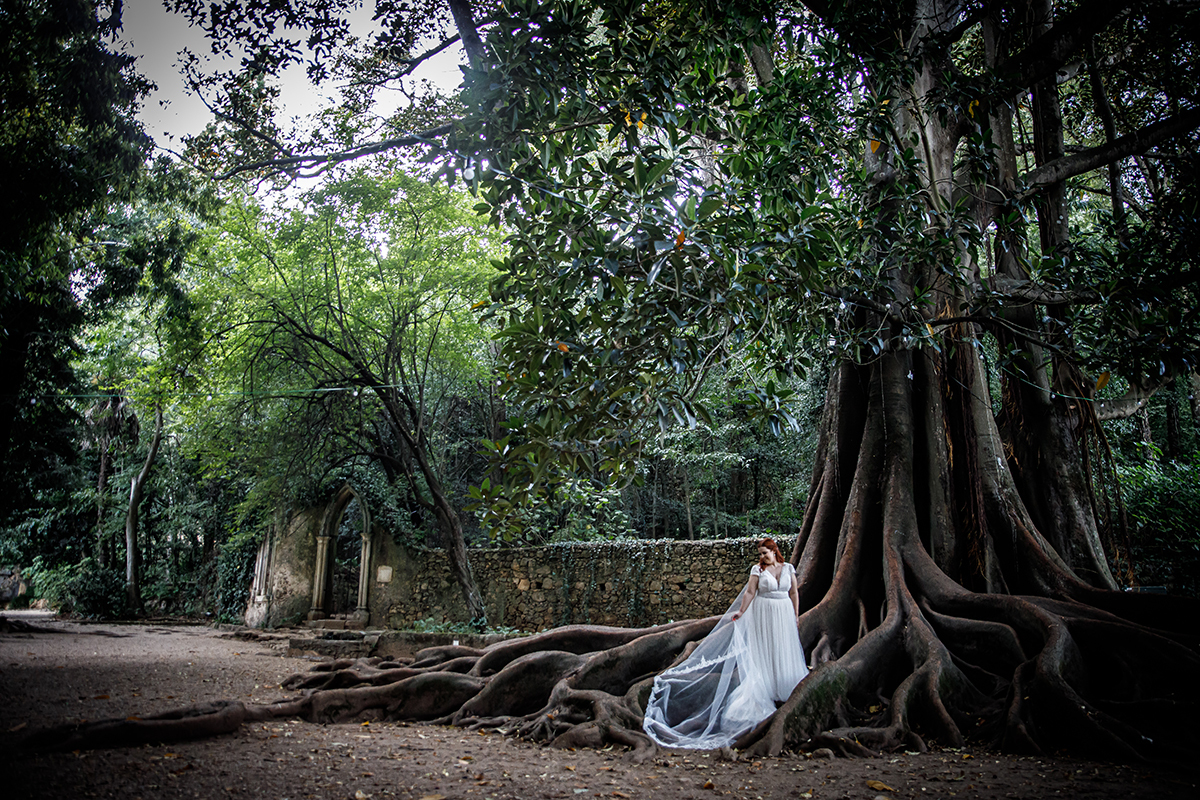 Mónica surge entre as raízes da árvore centenária. De corpo inteiro, olha para a câmara com um sorriso leve e postura graciosa. A preto e branco, o cenário ganha uma aura mágica, onde o tempo parece suspenso.