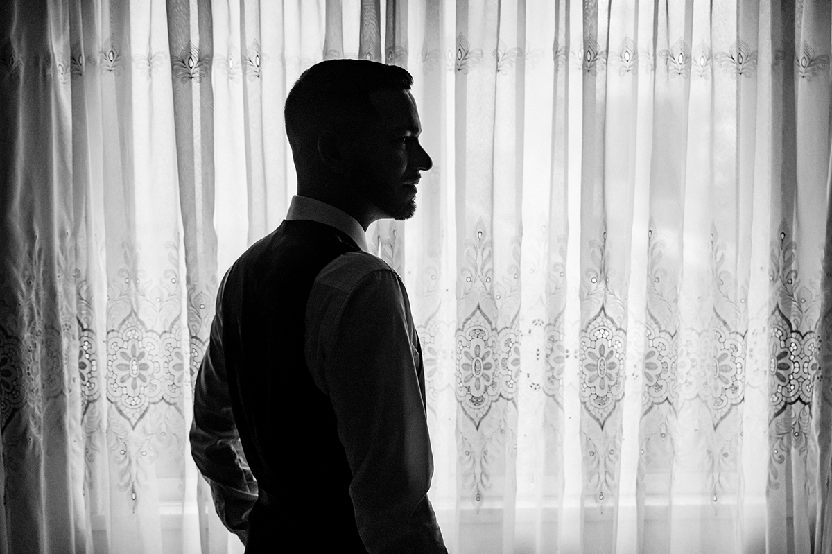 Envolto pela luz suave que atravessa a cortina branca, César surge em silhueta, um homem prestes a começar um novo capítulo.