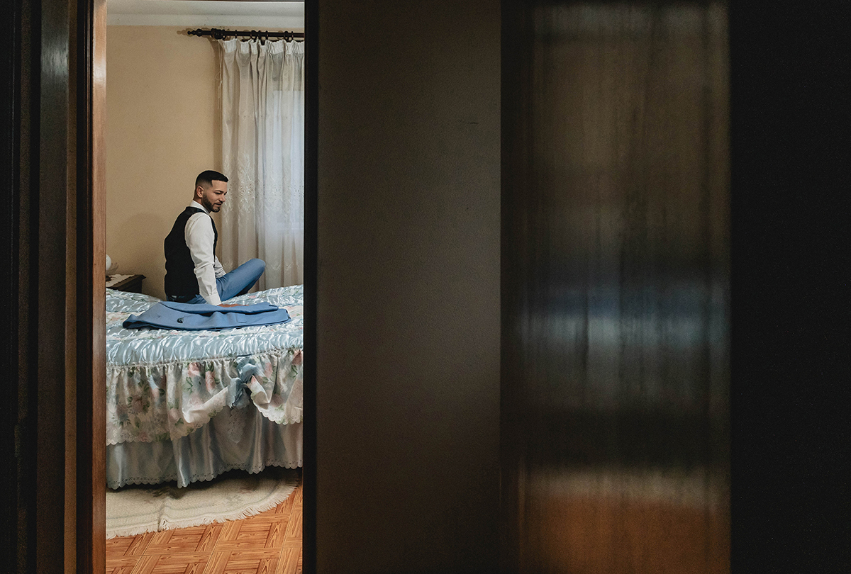 Sentado na cama, num momento de introspecção, César observa o seu fato azul impecável, símbolo da elegância e da importância daquele instante.