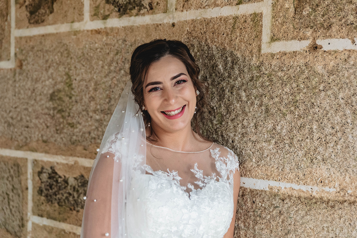 Contra a parede de pedra, o sorriso da noiva brilha, e as pérolas do véu completam a aura mágica da sua presença.
