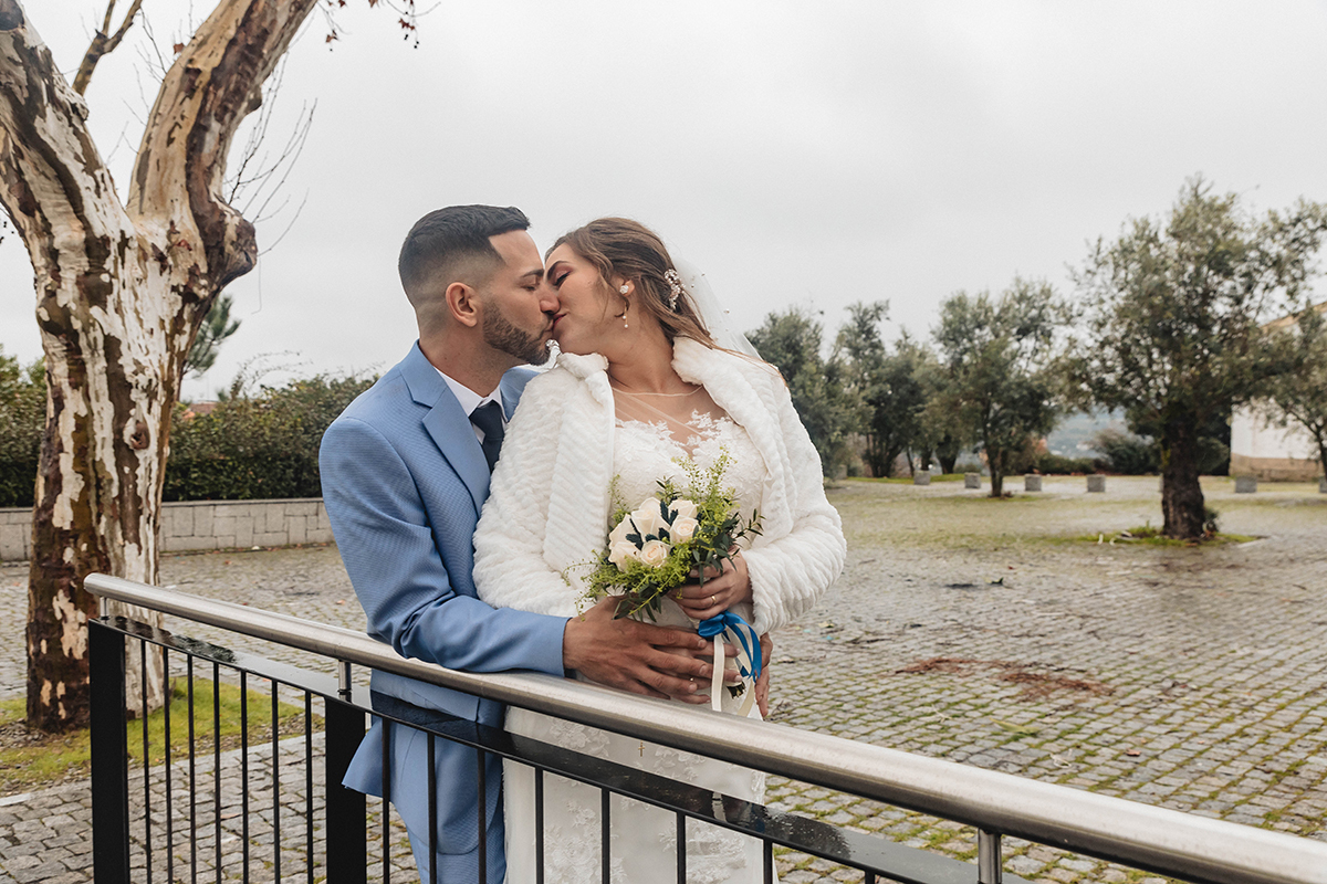 Junto ao corrimão, César abraça Sandra por trás e beija-a com ternura, pousando a mão sobre o seu ventre, um gesto de proteção e carinho.