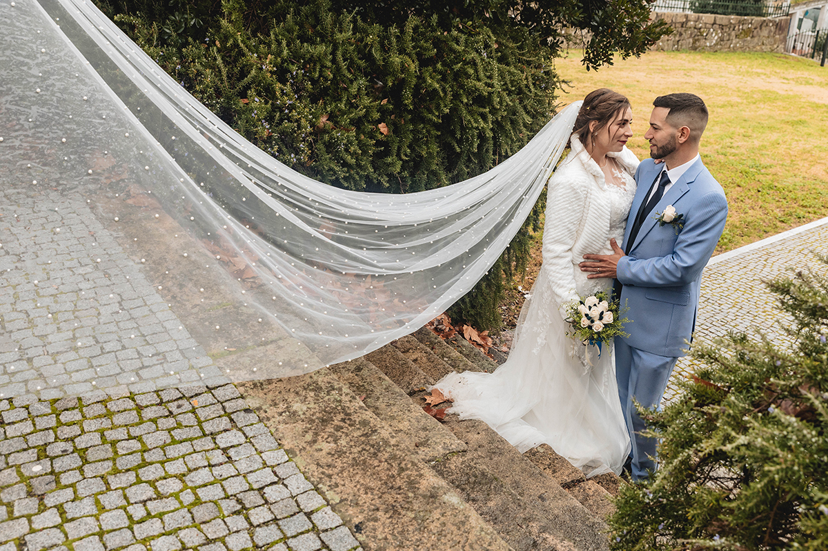 No topo da escadaria, o véu da noiva escorre em ondas brancas, enquanto os dois se encaram num momento cheio de promessas.