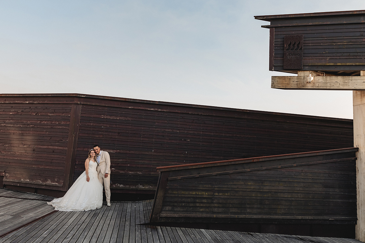 Do lado direito da imagem, o casal aparece com as cabeças encostadas, felizes. Atrás deles, uma estrutura de madeira com linhas modernas e toques vintage, em contraste com o céu azul deslumbrante.