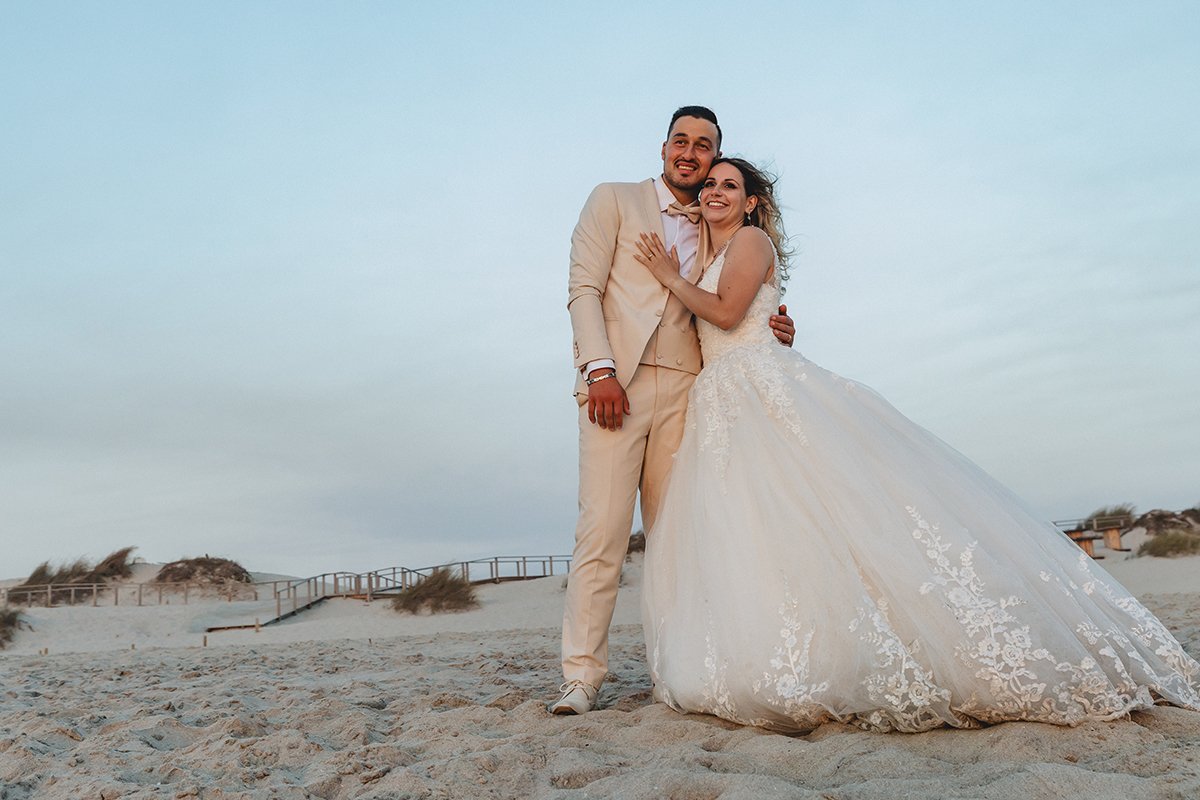 Agora à beira-mar, os dois sorriem ao olhar o oceano. Sabrina pousa a mão no peito do Jorge, enquanto ele a abraça por trás. Um céu azul e passadiços ao fundo completam a paisagem.