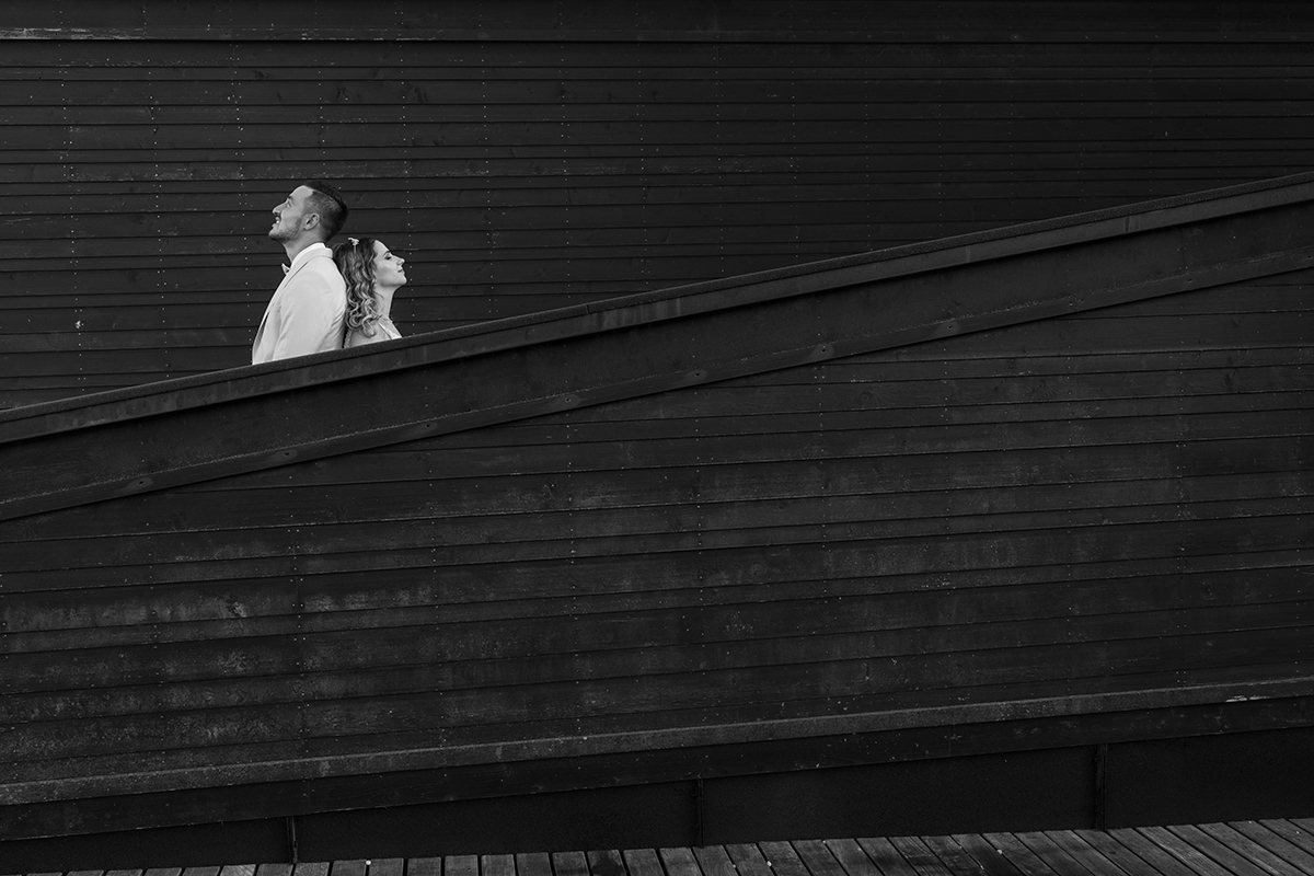 Uma imagem a preto e branco revela apenas os ombros de Sabrina e o peito de Jorge por detrás de um muro de madeira. Estão de costas um para o outro, a olhar o céu, num momento sereno e reflexivo. sessão trash the dress