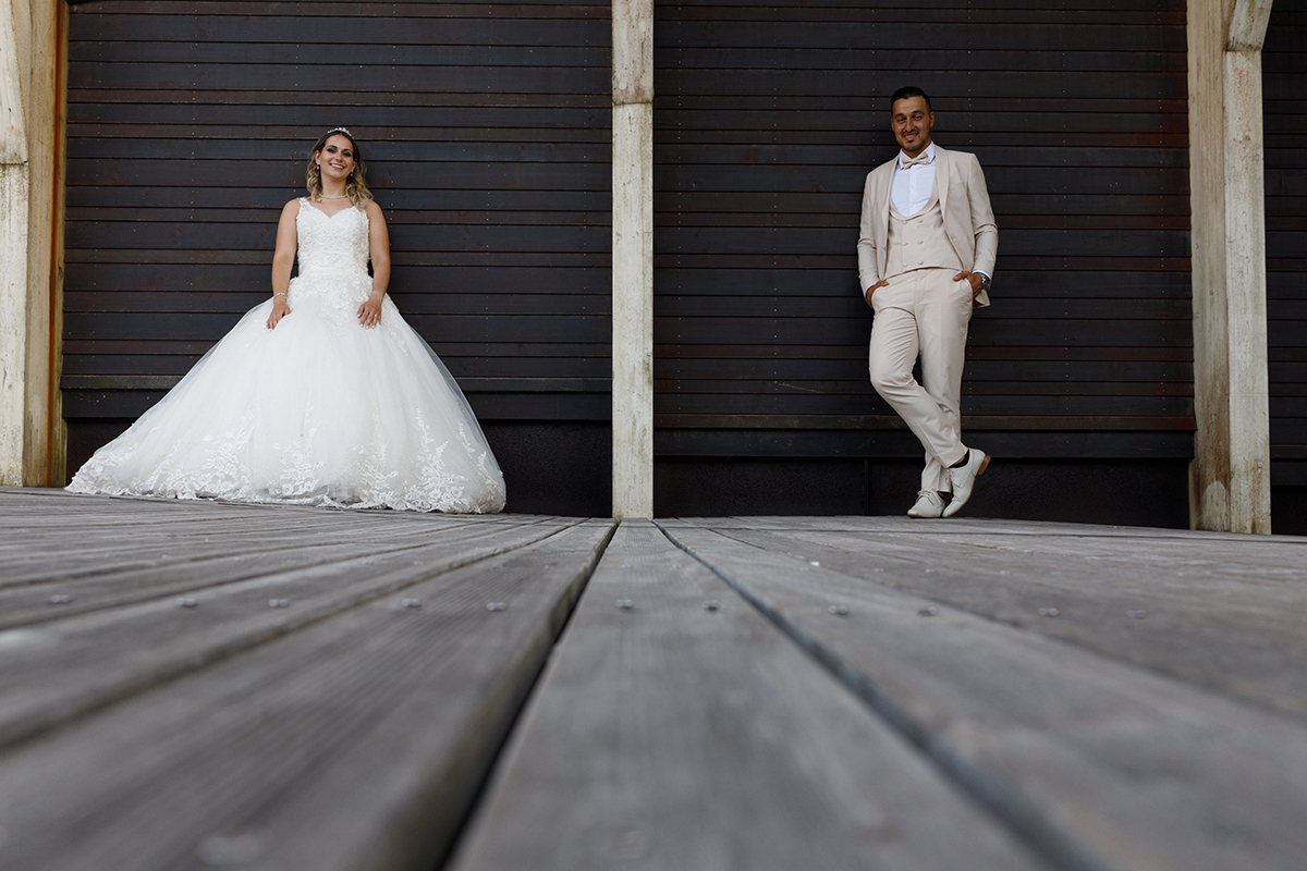 Fotografia tirada a partir do chão, captando os passadiços de madeira. Ao fundo, separados por um pilar mas junto à parede de um edifício, estão Sabrina e Jorge, sorridentes para a câmara.