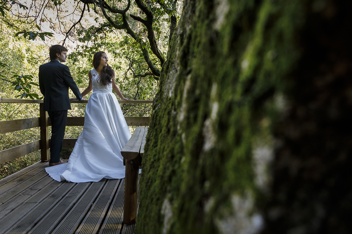 Um tronco coberto de musgo emoldura esta imagem serena. André e Diana, encostados ao corrimão, partilham um olhar sério sobre a paisagem. A Natureza observa-os em silêncio.