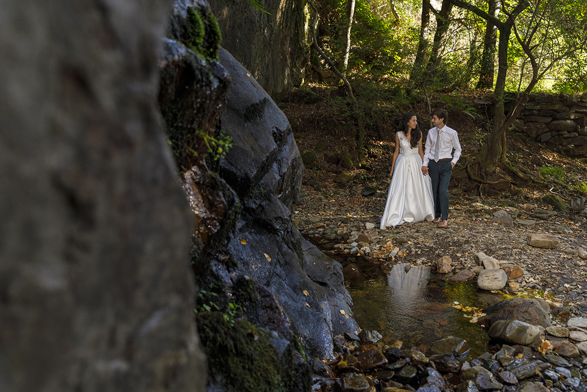 Uma imagem com profundidade: um rochedo desfocado em primeiro plano, outro mais definido, e ao fundo os noivos de mãos dadas ao pé de um pequeno lago, encarando-se com carinho e introspecção.