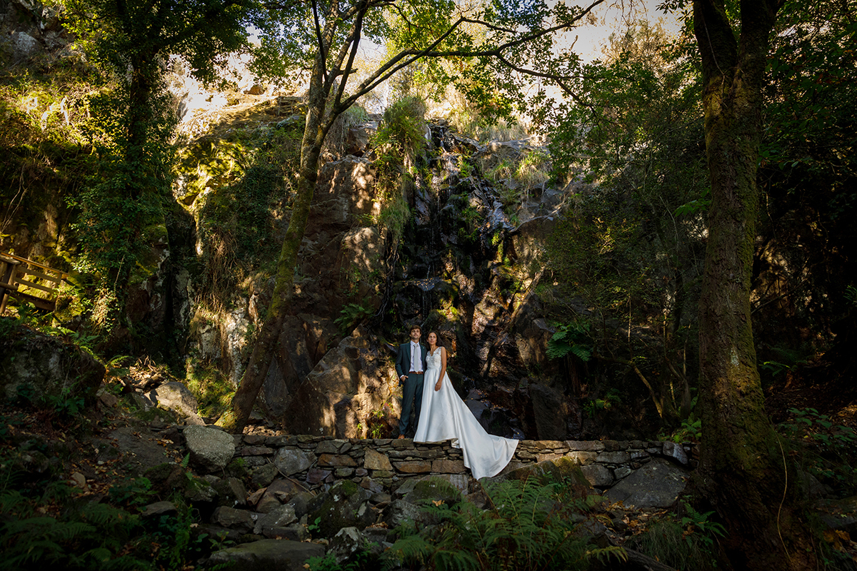 Em cima de um muro de pedra, lado a lado e sérios, os dois olham para a frente. Atrás deles, uma cascata fluente reforça a energia natural e a força do momento.