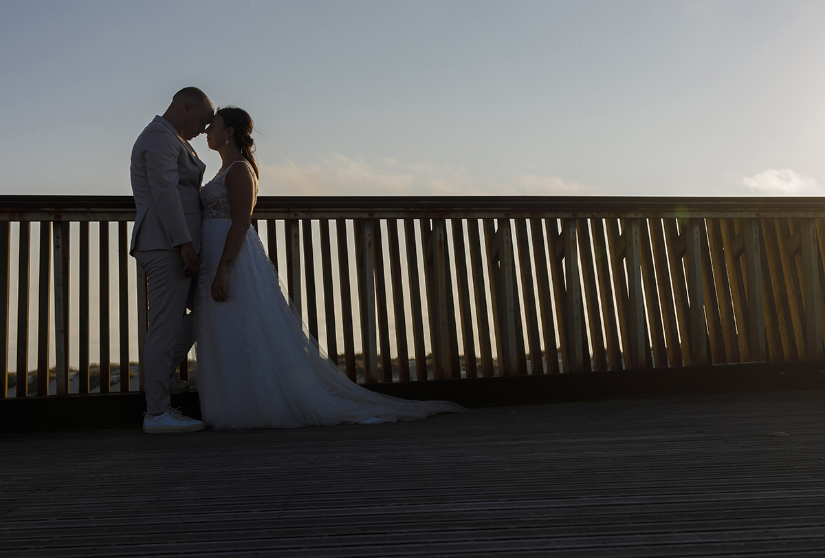 Uma imagem captada de baixo mostra os dois prestes a beijar-se, com as caras encostadas. O céu azul e amarelado contrasta com o muro de madeira perfurado que surge atrás.