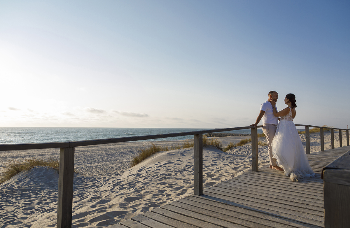 No passadiço, encostados ao lado oposto, Bruno está com as mãos atrás das costas, e Patrícia, à sua frente, toca-lhe suavemente na cara. Ao fundo, um cenário magnífico da praia.