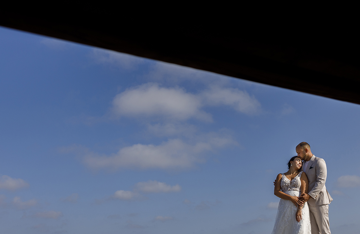 No lado direito da imagem, Bruno abraça Patrícia por trás, com as mãos nos seus braços. Ela está encostada ao peito dele e recebe um beijo carinhoso na testa.