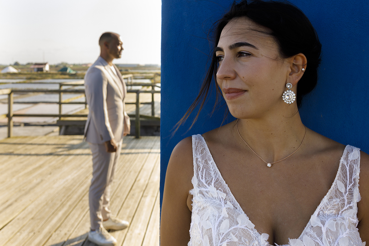 Patrícia aparece encostada a uma parede azul, fotografada do peito para cima, enquanto Bruno está mais atrás, de corpo inteiro, a olhar em frente, com a luz do sol a incidir sobre ele.