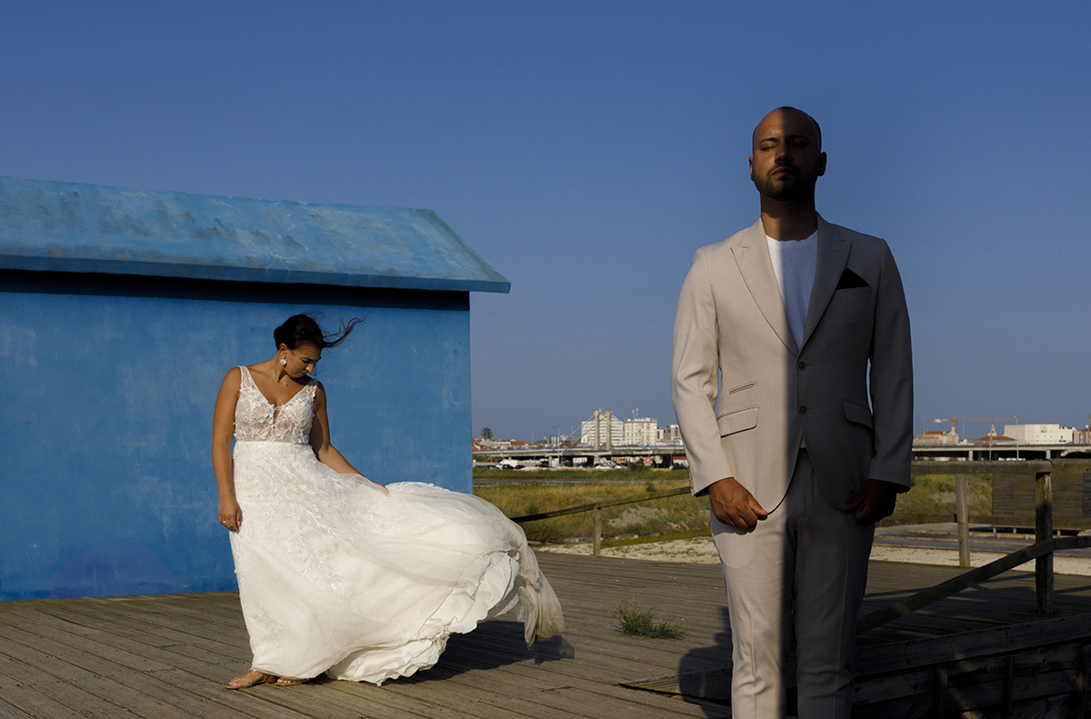 Bruno surge mais próximo da lente, no lado direito, com metade do corpo iluminado e a outra em sombra. Atrás, à esquerda, Patrícia observa o vestido que esvoaça com o vento.