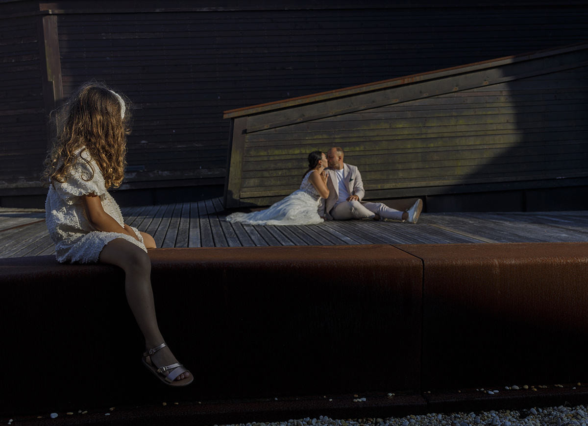 Sentados sobre uma base de madeira, com uma parede inclinada atrás, Patrícia e Bruno beijam-se no centro da imagem. À frente, a sua filha está sentada no parapeito a olhar para eles, criando um retrato de família ternurento.