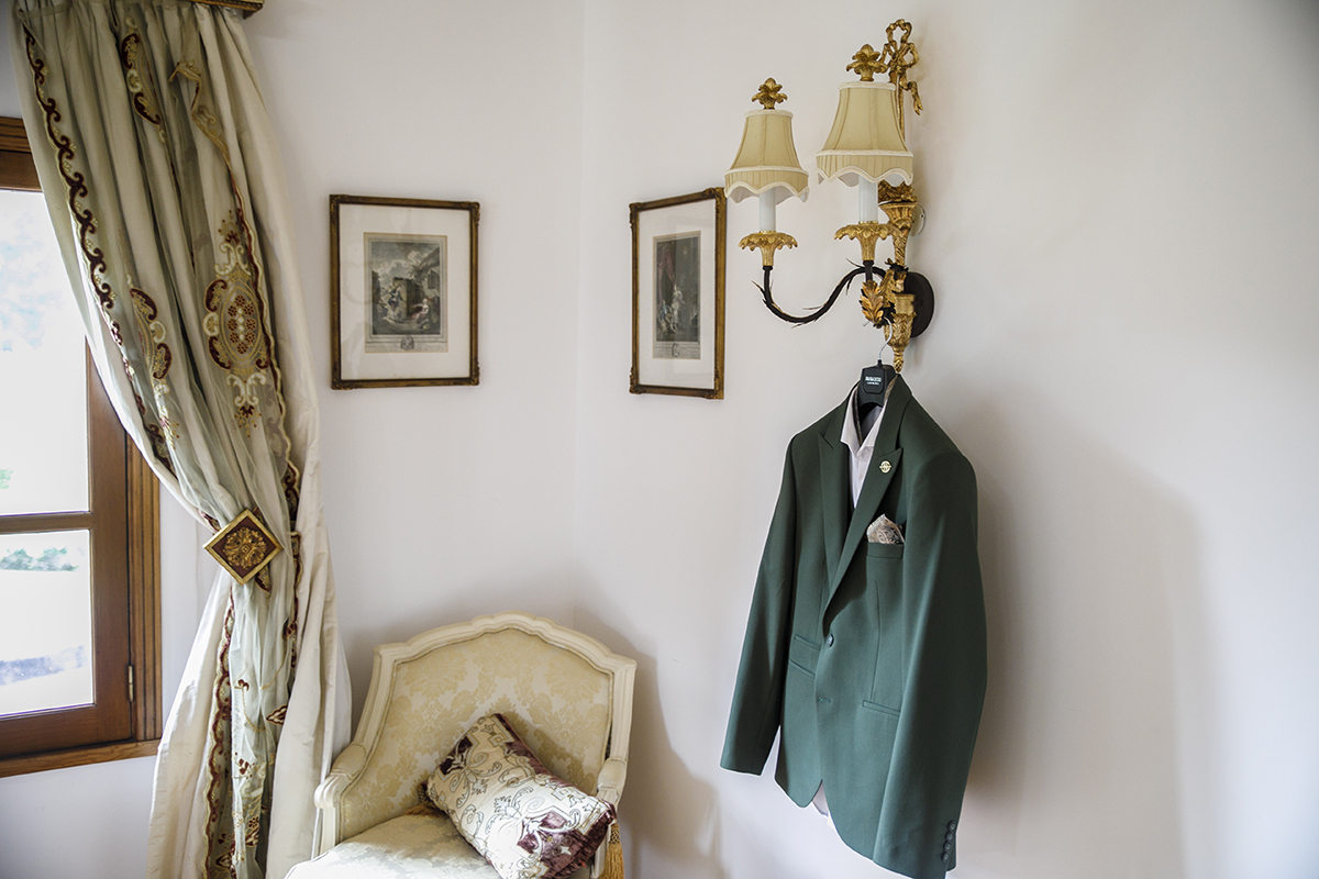 O fato do noivo, Furkan, está pendurado num candeeiro antigo, numa sala de parede branca, com uma poltrona bege e cortinas junto à janela.