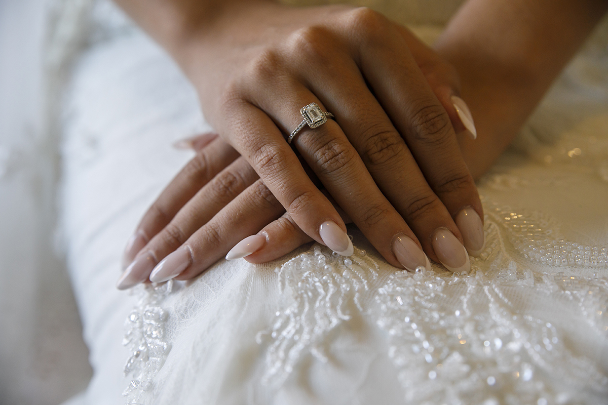 Num plano aproximado, destacam-se as suas mãos cruzadas sobre o vestido e o anel.