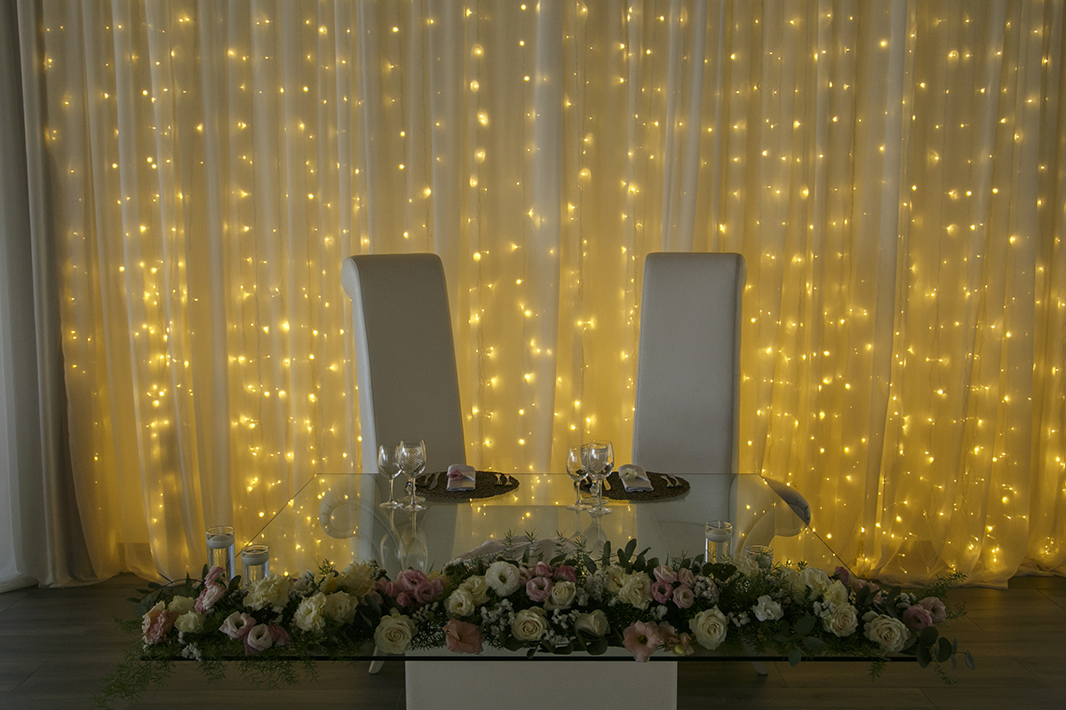 Detalhe da mesa decorada com flores e as cortinas com luzes amarelas, cheia de charme.