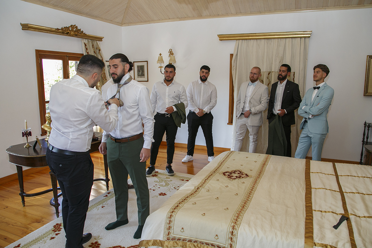 O padrinho principal ajusta a gravata de Furkan, enquanto os restantes amigos observam ao fundo, sorridentes.