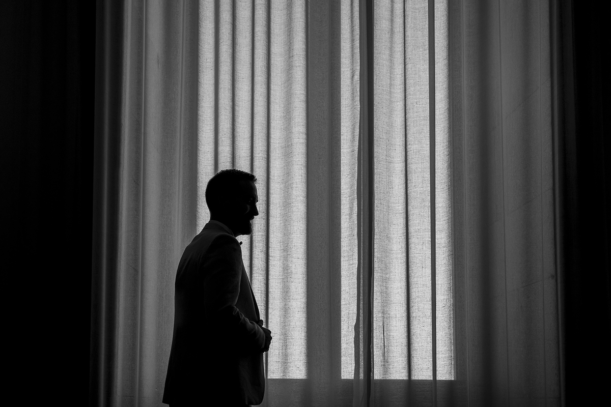 fotografos casamento viseu. Uma silhueta diante de uma grande janela com cortinas translúcidas. Mathieu, de perfil, observa em silêncio. A luz abraça-o, como se a própria manhã abençoasse o seu caminho.