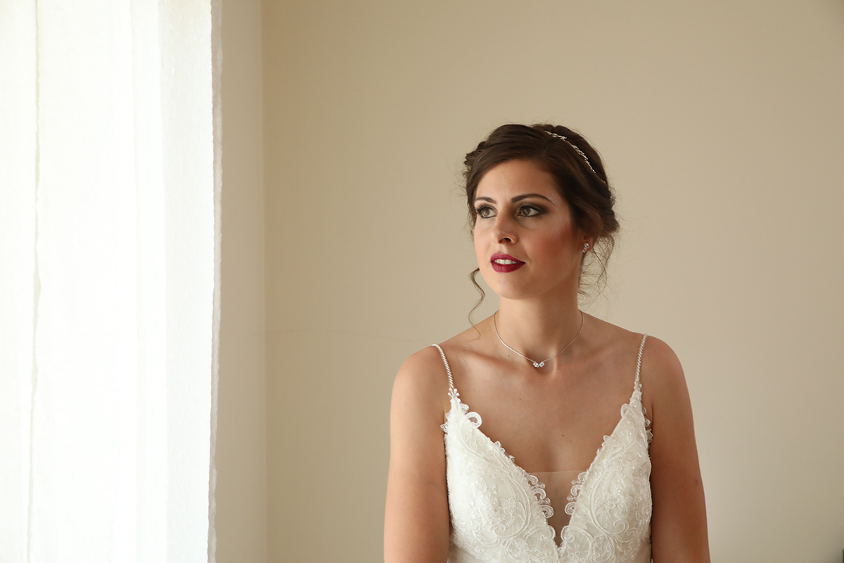 Marta, do peito para cima, observa a janela com olhar fixo e pensativo. A luz suave e a parede branca compõem um cenário puro, onde se sente a emoção prestes a florescer.