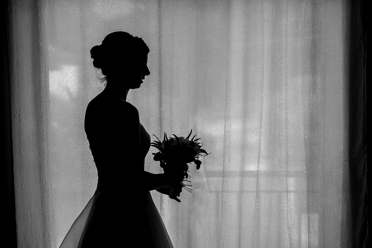Numa fotografia em silhueta, Marta surge delicada junto à janela. De lado, com o bouquet na mão, a sua figura ganha contornos quase mágicos sob a luz difusa.