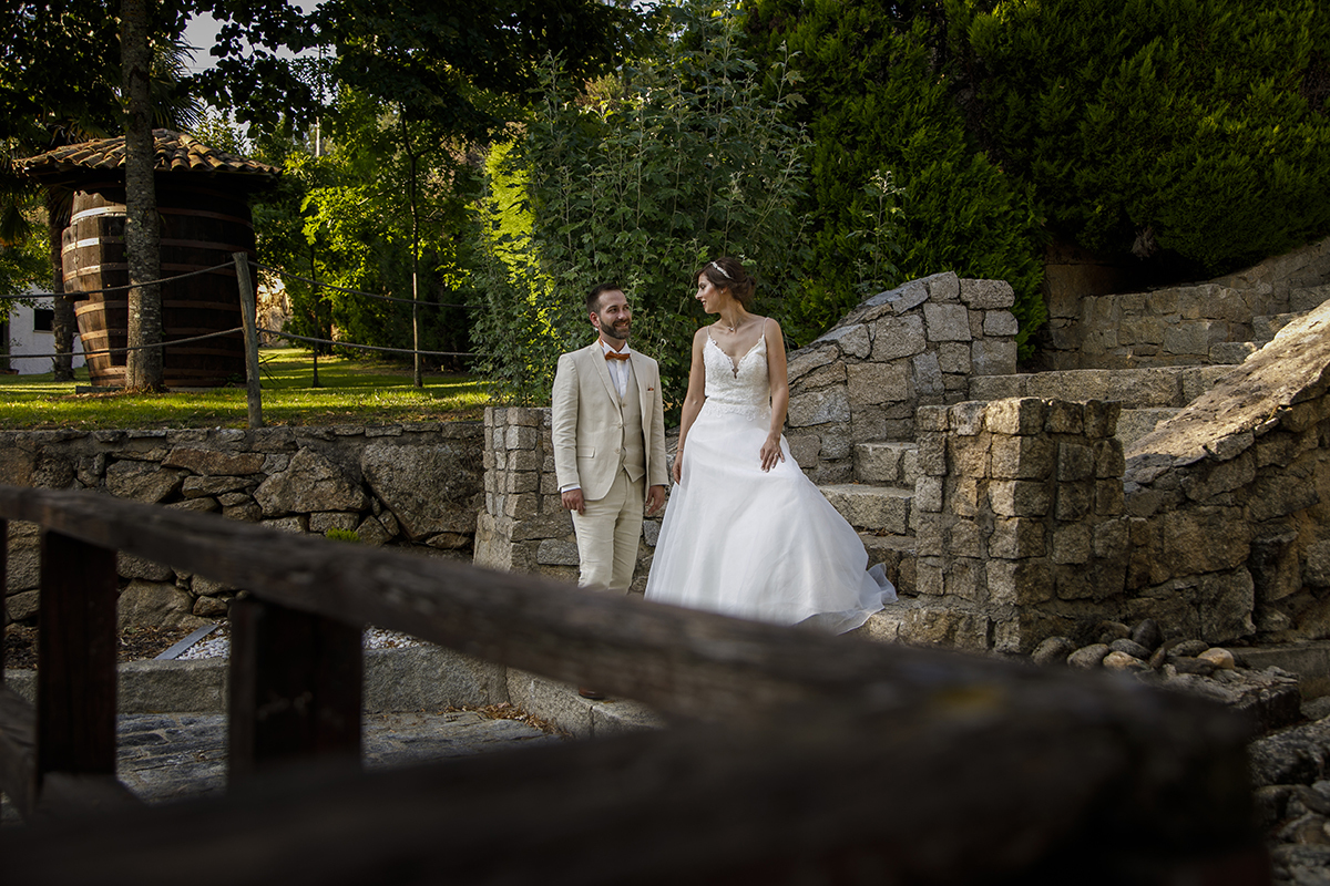 Descem por uma escadaria entre verdes e pedras, de mãos dadas, sorridentes. A madeira desfocada em primeiro plano dá profundidade ao momento, como se o mundo parasse para os ver passar.