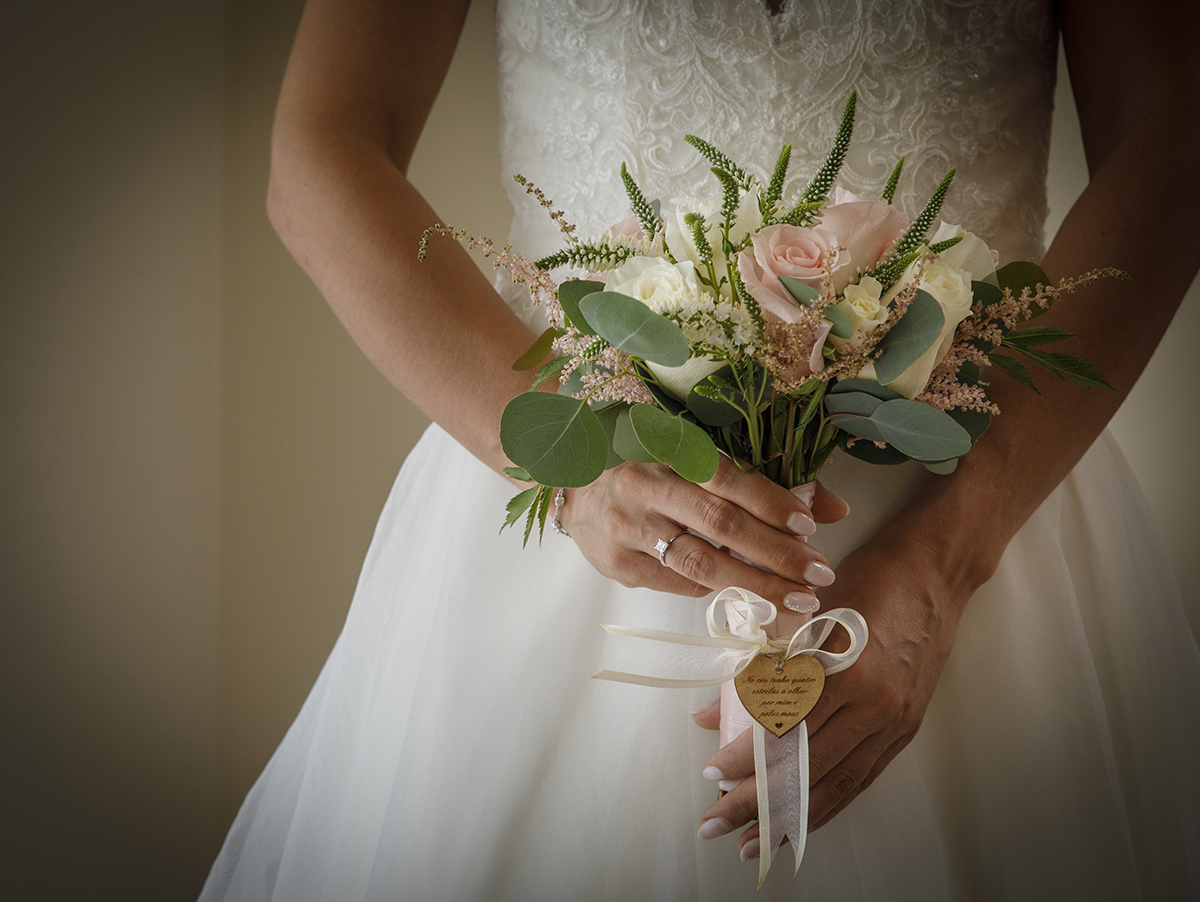 Um retrato com vinheta realça as mãos delicadas de Marta segurando o seu bouquet. Flores rosas e brancas, um lacinho e um pequeno coração contam uma história de amor, carinho e beleza.