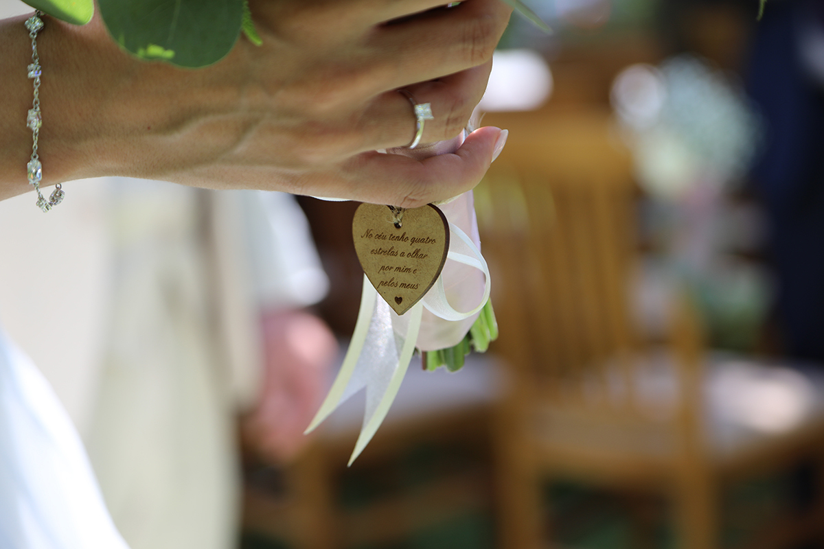 Um plano de detalhe delicado. As mãos de Marta seguram o bouquet com o laço e o coração em destaque. Um símbolo de amor captado com sensibilidade.