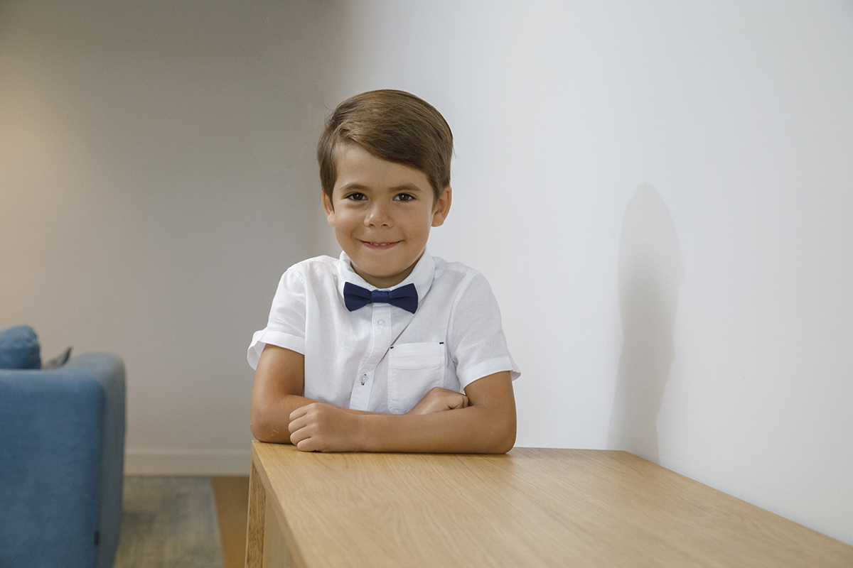 De braços cruzados sobre um móvel de madeira, Ethan olha diretamente para a câmara com um brilho especial no olhar. Ao fundo, um toque de azul do sofá complementa esta imagem alegre e intimista.