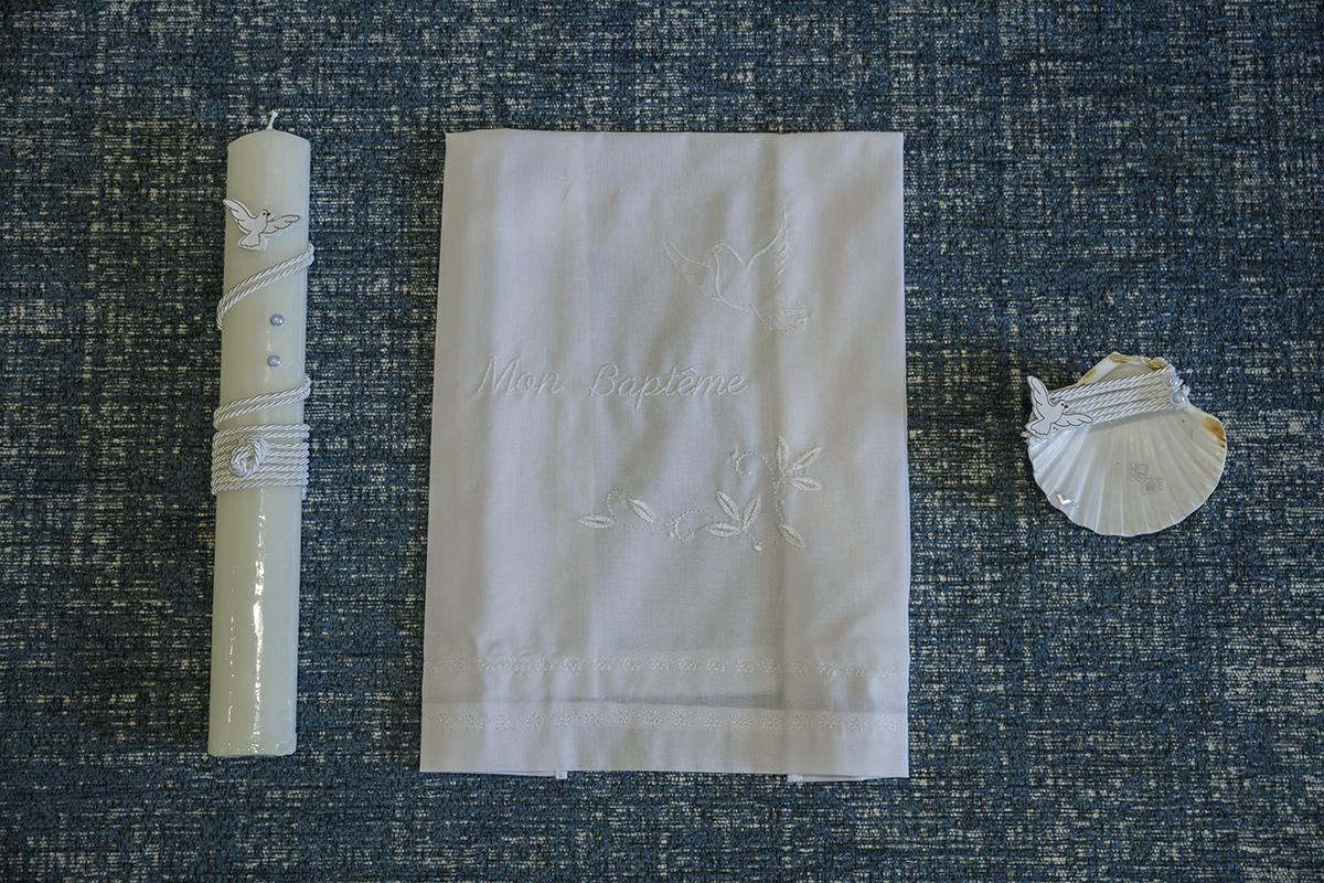 Sobre um fundo azul suave, destacam-se três símbolos sagrados: a vela, o pano branco e a concha do batismo, elementos que representam fé, pureza e renovação espiritual neste dia tão especial para o pequeno Ethan.