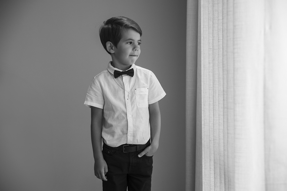 Fotografos de batizado. Esta imagem a preto e branco transmite introspeção. Ethan olha para a janela, morde o lábio e mantém uma mão no bolso. A luz suave das cortinas brancas destaca a expressão pensativa do pequeno modelo.