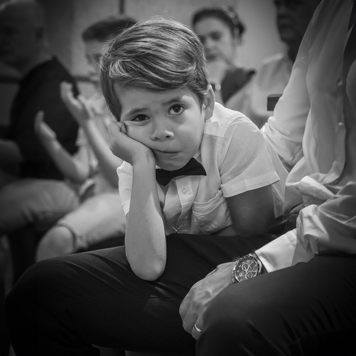 Uma imagem a preto e branco com forte carga emocional: Ethan apoia os braços na perna do pai enquanto sente o toque carinhoso da mãe no seu rosto. O seu olhar revela pensamentos profundos, num registo cheio de ternura e ligação.