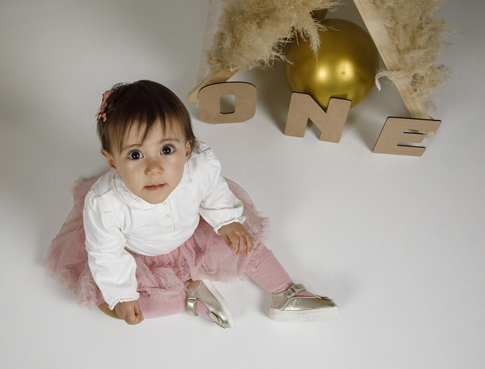 Fotografia primeiro aniversario. Numa vista picada, Matilde está no lado esquerdo da imagem, sentada no chão branco, com as mãos na saia rosa e a olhar para cima. Ao fundo, destaca-se uma cabaninha com balão dourado e as letras “ONE”.