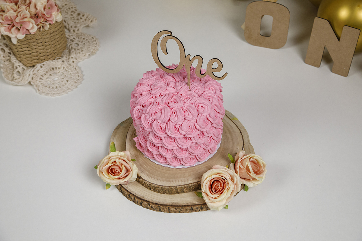 Neste plano picado, vemos o bolo rosa da Matilde ao centro, com rosinhas e a palavra “ONE”. À direita, balão dourado; à esquerda, cesto de rosas. Um cenário encantador que rouba todas as atenções.