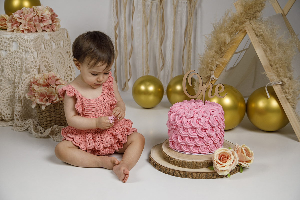 Sentada junto ao seu bolo, Matilde já tem pedacinhos nas mãos e observa-as com uma expressão séria. A doçura do momento é realçada pelo contraste entre o bolo rosa e o chão branco.