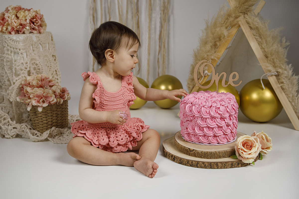 Sentada no mesmo cenário, Matilde olha com curiosidade para o seu bolo rosa, já com a mãozinha pousada nele. Um momento cheio de ternura, captado com muita sensibilidade.