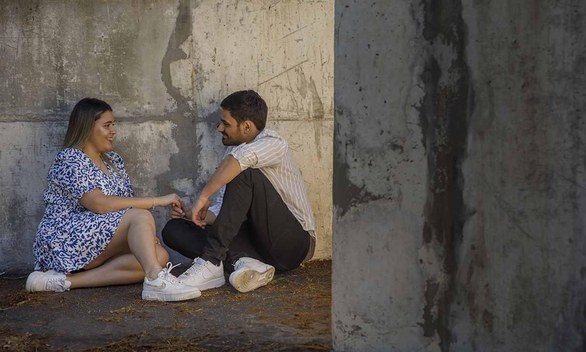 Sentados no chão, pernas cruzadas, ela à esquerda, ele à direita, trocam olhares e sorrisos junto a uma parede, um cenário urbano captado numa curva que revela duas perspetivas. Fotografos casamento