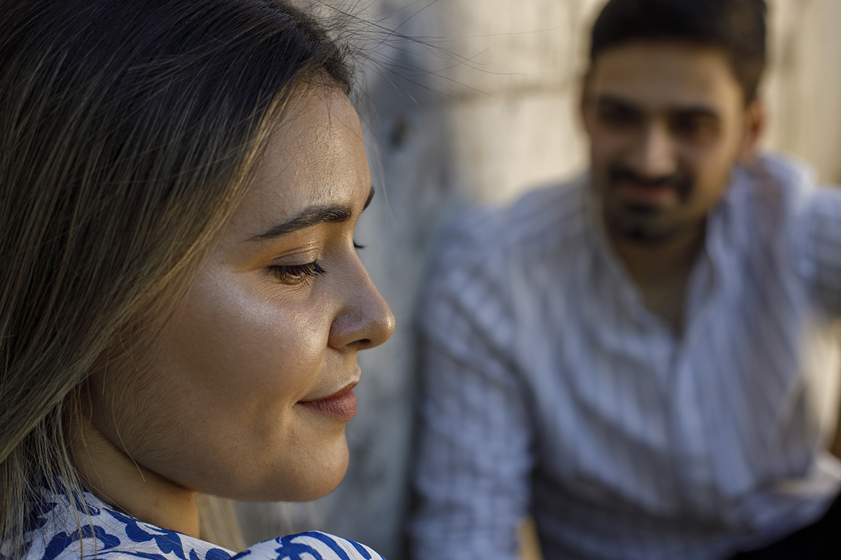 Um plano aproximado mostra Márcia nítida, a olhar para baixo com um sorriso terno. Ao fundo, Luís desfocado observa-a com expressão apaixonada.