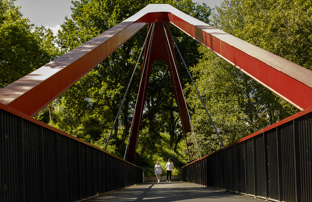 Uma bela paisagem rodeada de árvores emoldura a ponte vermelho-preta. Os dois caminham ao fundo, olhos nos olhos. Reportgem solteiros